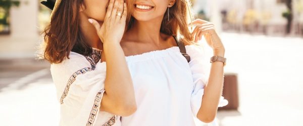 Fashion portrait of two young stylish hippie brunette and blond women in summer sunny day in white hipster clothes posing on the street background. Models share secrets, gossip.